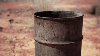 Naklejka premium A rusty metal barrel sits upright in a dry, barren area with red earth and sparse vegetation. The sunlight casts shadows, highlighting the textures of the weathered surface.