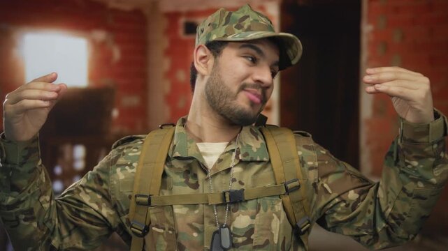 Man in camouflage uniform with backpack and dog tags raises both hands inside a brick building; duty focus discipline readiness.