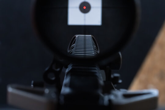 View through a rifle's optical scope (LPVO) with an illuminated red dot, aiming at a precision target.