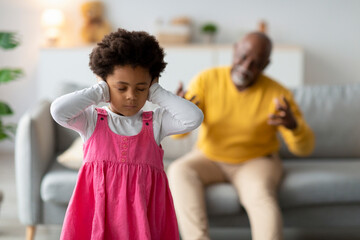 In a living room, a sad black girl covers her ears, not wanting to hear her grandpa's angry...