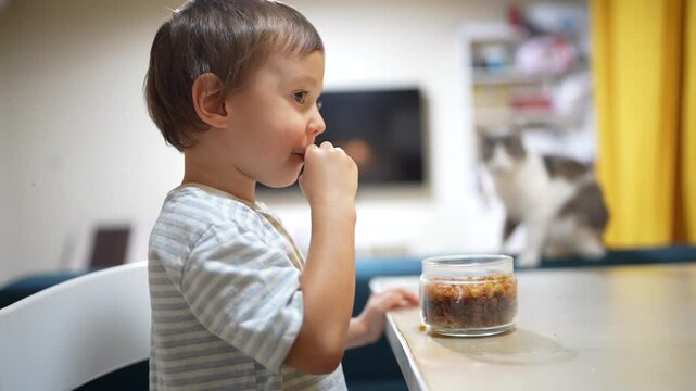 The young boy stands beside the kitchen table, intently looking at a jar filled with snacks. With curious fingers, the boy eagerly explores the tempting snacks inside the jar.