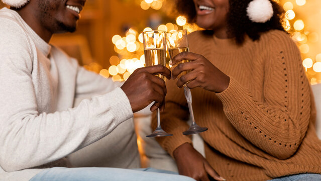Two happy millennial African American friends in Santa hats toast with champagne glasses in a festive living room. Glowing garlands create a warm holiday atmosphere as they celebrate together. - Powered by Adobe