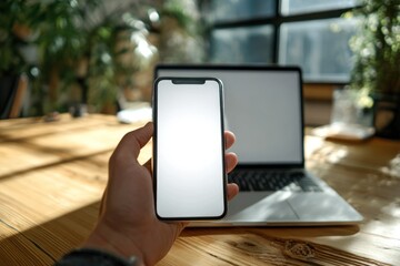 Hand holding smartphone with laptop on wooden desk indoors