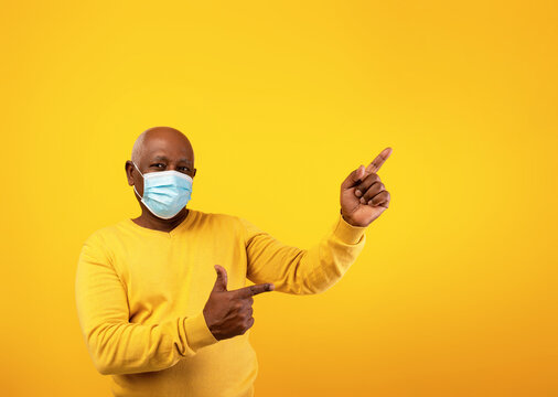 A senior black man wearing a protective face mask is pointing to the right against a bright orange background. He offers a space for text or advertisement, highlighting safety during the pandemic.