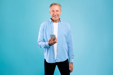 Cheerful senior man stands in a blue studio, using his smartphone. He looks at the camera with a...