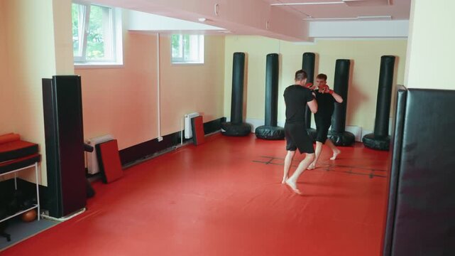 karate practitioners sparring in martial arts gym wearing gloves moving across red mat floor practicing offensive and defensive stance surrounded by punching bags during training session under