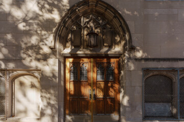 A pair of aged wooden doors set in a carved Gothic arch, framed by a stone façade. Dappled sunlight and shadows create a timeless, historic atmosphere of old architecture and quiet elegance.