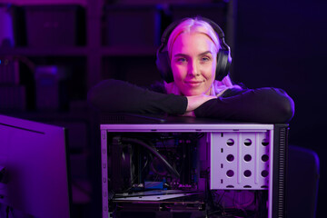Female gamer with headphones leans on an open gaming PC case under purple lighting, ready to upgrade hardware