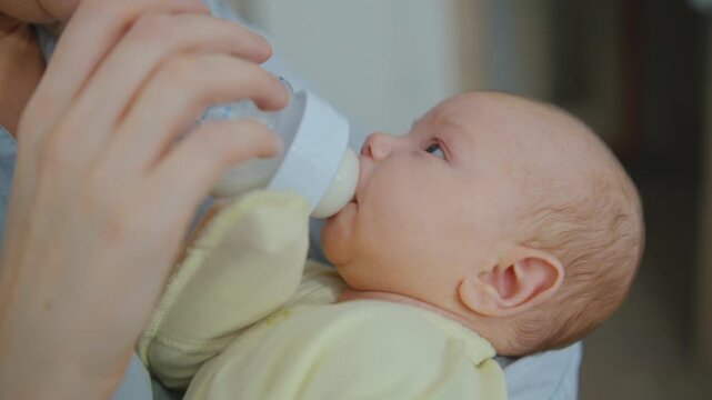 A mother is feeding a baby formula to a small infant from a bottle in a close-up. The baby is eating with appetite and looking at her