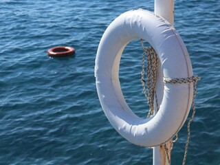 A white lifebuoy is secured to a pole beside calm blue water, with a distant red buoy visible. This nautical stock shot conveys safety, maritime themes, and travel vibes.