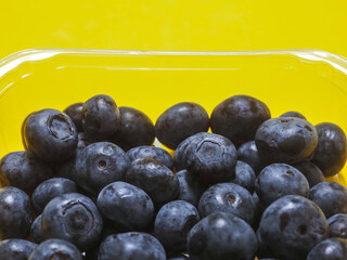 A plastic tray of blueberries sits on a bold yellow color background. The tray is clear and filled with fresh organic fruits ready for consumption. Healthy diet concept. Natural product.