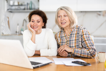Two cheerful women making video call by laptop sitting at the kitchen table