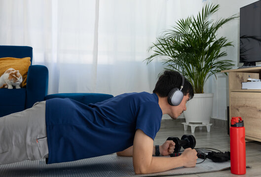 Man performing a plank exercise in his living room while listening to music with headphones, focused and concentrated. Concept of home workout, wellness, and active lifestyle - Powered by Adobe