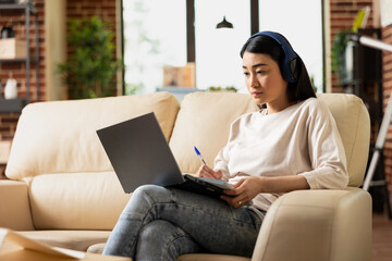 Professional asian woman sits on couch with notepad and laptop, reviewing notes for business strategy. Focused female individual working from home as virtual assistant, writing checklist for employer.