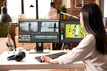 Female videographer working from apartment, assembling film footage on desktop monitors. Focused...
