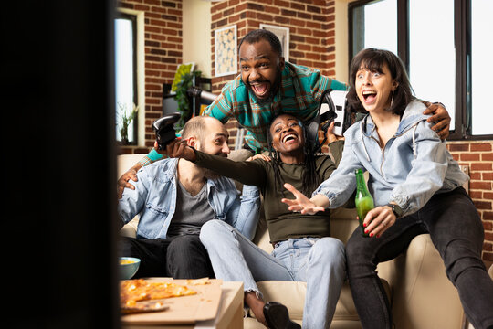 African american and caucasian friends enjoy VR games in brick wall apartment. Black woman celebrates gaming victory as diverse group cheering with drinks in hand at joyful indoor hangout. - Powered by Adobe