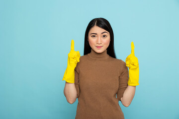 Asian woman with yellow rubber gloves pointing upwards with both index fingers, standing against blue background. Female janitor gesturing towards ceiling, directing focus to copy space above.