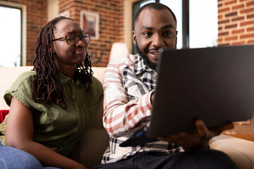 Black man gestures to laptop screen while discussing vacation ideas with girlfriend on sofa. Young couple reviewing presentation together, balancing work talk and personal planning at home.