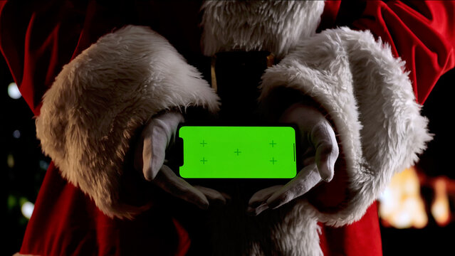 Santa Claus hands in a red suit and white gloves hold a smartphone with a green screen chroma key. A warm festive atmosphere for digital or themed mockups.