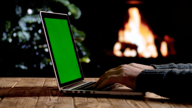 A man works on a laptop with a green screen chroma key background in front of a fireplace and a Christmas tree. A warm festive atmosphere for digital or themed layouts.