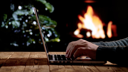 A man works on a laptop in front of a fireplace and a Christmas tree. Freelancing, remote work, or studying at home during the holidays. A warm festive atmosphere for New Year or Christmas.