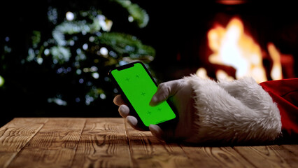 Santa Claus hand in a white glove holds a smartphone with a green screen chroma key background in front of a fireplace and a Christmas tree. A warm festive atmosphere for digital or themed mockups.