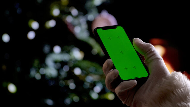 Santa Claus hand in a white glove holds a smartphone with a green screen chroma key background in front of a fireplace and a Christmas tree. A warm festive atmosphere for digital or themed mockups.