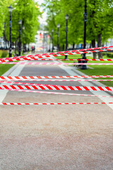 Urban park walkway blocked with red and white warning tape restricting access to public area. Concept of closure, restriction, and emergency safety in city environment