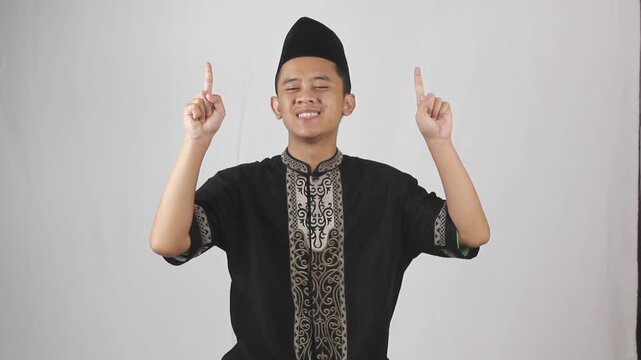 Cheerful Muslim man wearing black Koko clothes pointing his finger up against a white background - Powered by Adobe
