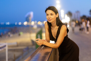 Fototapeta premium Relaxed young girl stands by the seaside railing under warm evening lights in Barceloneta promenade