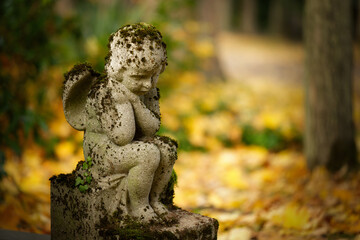 Obraz premium A deeply sorrowful, mossy stone cherub statue sits head bowed in contemplation amidst the vibrant golden decay of fallen autumn leaves