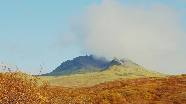 A scenic view of Kristinartindar mountain in Skaftafell National Park, Iceland, with a cloud partially obscuring the peak. The foreground is filled with vibrant orange and yellow autumn foliage under 