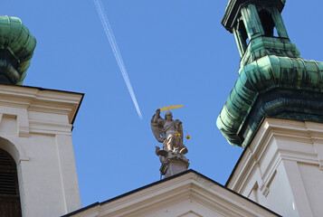 An airplane and its white trail passing the Statue of Archangel Michael atop the Church of St. Michael the Archangel (Czech: Kostel svateho Michaela archandela) in Brno, Czechia