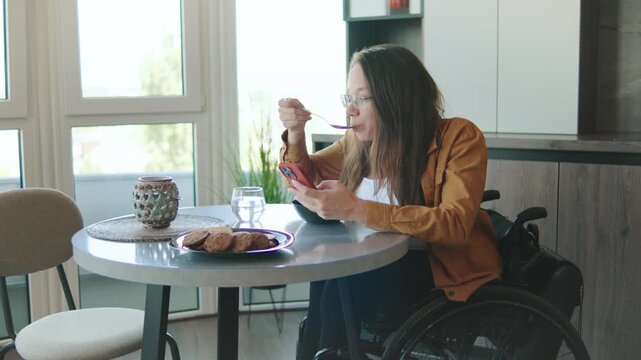 Woman in a wheelchair enjoying breakfast while using a smartphone at home, showcasing inclusion and daily life with a focus on mobility and independence