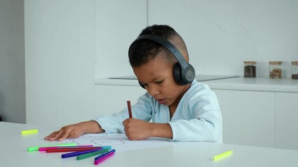 A young African American boy is focused on his drawing at home. He uses colorful markers to create art while wearing headphones, enjoying a moment of creativity and solitude.