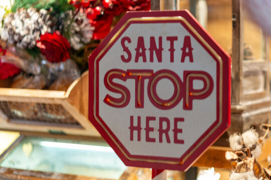 A bright red and white road stop sign with red text inviting Santa Claus to stop here. The colourful, cheerful hanging sign is part of the Christmas decor in a shop. There are flowers in a basket. 