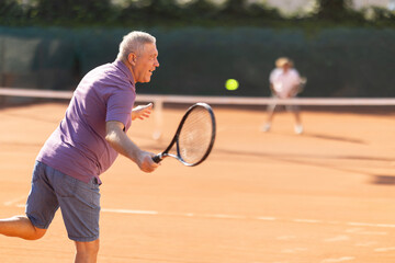 Elderly man in sportswear plays tennis on a tennis court in the summer. Pensioner hits a tennis ball with a racket