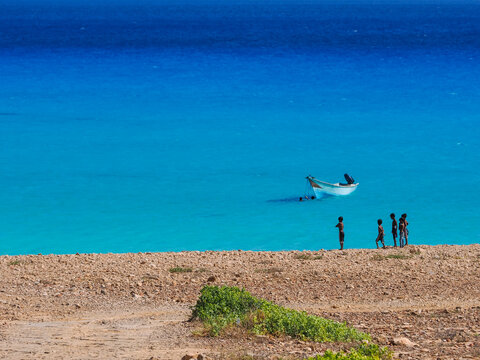 Group of children on beach looking at fishing boat on turquoise water, Socotra Island, Yemen