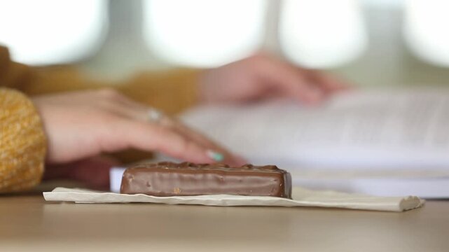 A student is focused on reading a textbook, then pauses to pick up and eat a chocolate bar placed on the table, taking a study break.