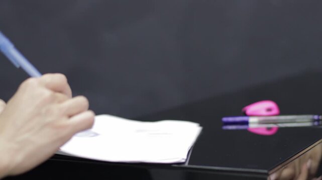 A professional's hand writes with a blue pen on white paper resting on a shiny black desk. Ideal for depicting office work, academic tests, note-taking, or contract signing.