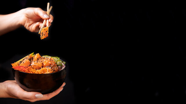 Detalhe de m&atilde;o segurando um hashi com um cubo de salm&atilde;o cr&uacute; e fresco com gergelim branco e preto e bowl de poke . Ilumina&ccedil;&atilde;o de est&uacute;dio e fundo preto, publicidade de gastronomia 