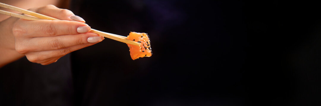 Detalhe de m&atilde;o segurando um hashi com um cobo de salm&atilde;o cr&uacute; e fresco com gergelim branco e preto. Ilumina&ccedil;&atilde;o de est&uacute;dio e fundo preto, publicidade de gastrornomia 