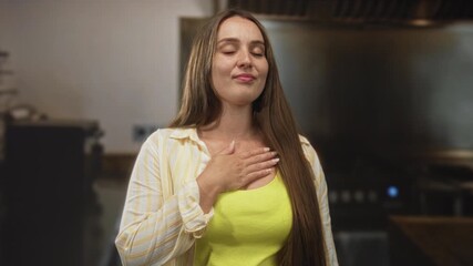 Woman with hand on chest in kitchen building, wearing yellow shirt and striped overshirt, eyes closed and slight smile; quiet gratitude.