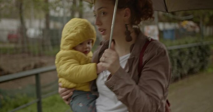 A mother holds her baby close while walking in the rain under an umbrella. They are near an enclosure, and a stroller is also visible.