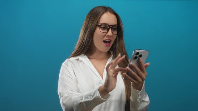 Young caucasian woman wearing glasses and a white shirt tapping smartphone screen with hands in blue studio; surprise reaction.