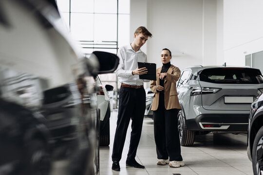 Young man in white shirt and woman in brown blazer discussing car features in a modern dealership, showcasing customer service and automotive sales experience
