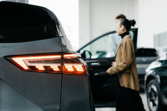 Woman in stylish coat is walking towards a modern car in a dealership, showcasing the sleek design and vibrant lighting of the automotive environment