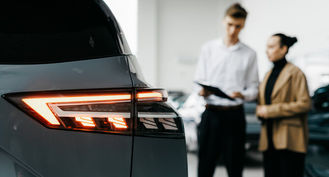 Car dealership scene featuring a man and woman discussing vehicle features, with a close-up of a car's taillight, showcasing modern automotive design and sales interaction