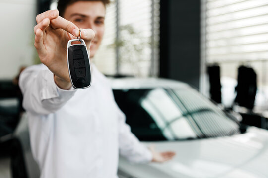 Young man in a white shirt proudly holding car keys in a modern dealership, showcasing the excitement of purchasing a new vehicle with bright showroom ambiance