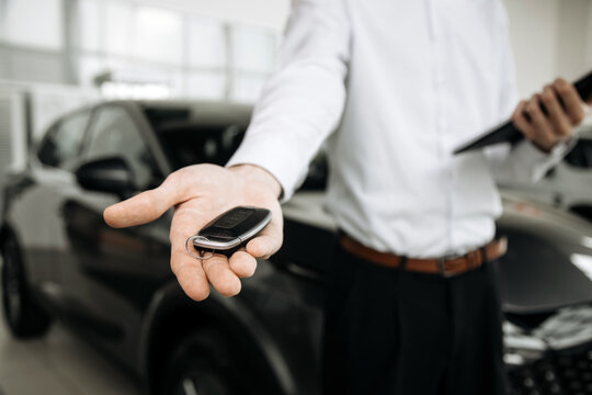 Professional man in formal attire holds car key fob in hand, showcasing vehicle purchase process at modern car dealership with sleek cars in background